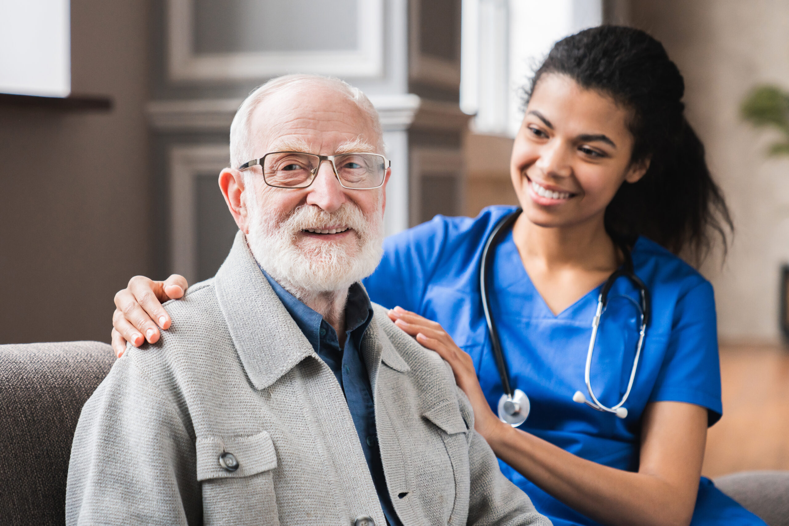 Young nurse in blue coat hugging old 80s man smiling looking at camera. Portrait of satisfied elderly patient and his caregiver. Medical care of older generation people, geriatrics medicine, nursing concept